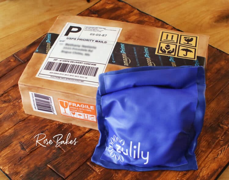Cake shaped like and amazon cardboard shipping box and a blue zulily package.  The cake is sitting on a cake board that looks like wood made from fondant and hand painting.