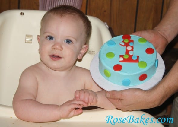 A picture of Asher (Rose Bake's son) smiling in a highchair with blue red and green polka dots 1st Birthday Smash Cake being held next to his head.