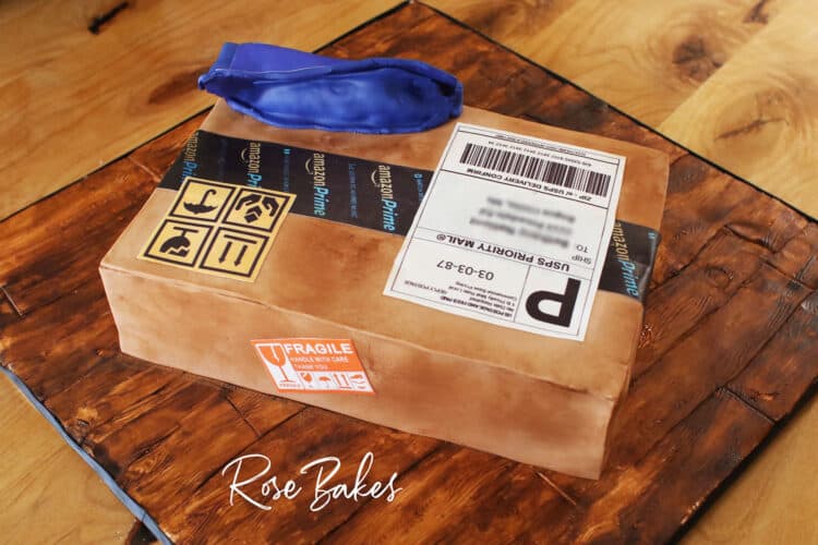 Back view of a cake shaped like and amazon cardboard shipping box and a blue zulily package.  The cake is sitting on a cake board that looks like wood made from fondant and hand painting.