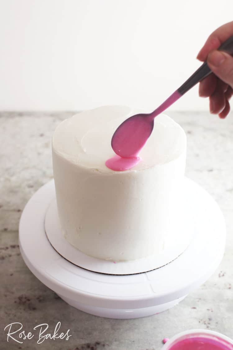 A spoon of pink canned frosting being added to the edge of a chilled white cake on a cake stand