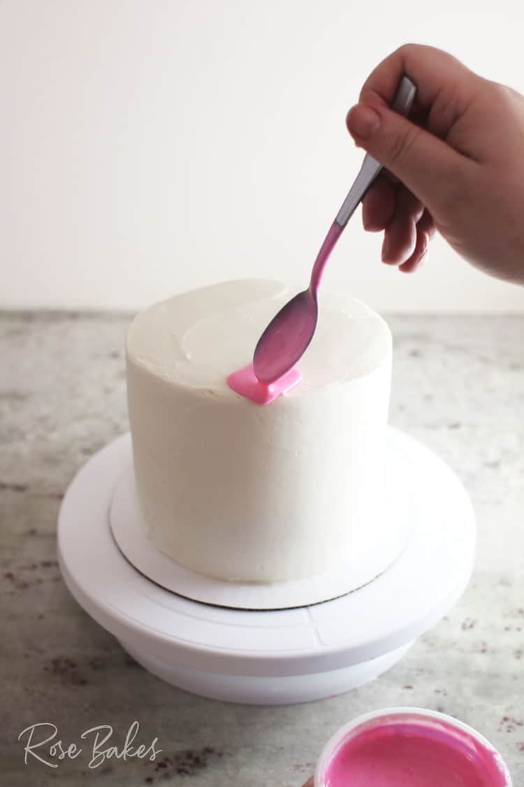 A spoon of pink canned frosting being added to the edge of a chilled white cake to create drip. the cake is on a cake stand