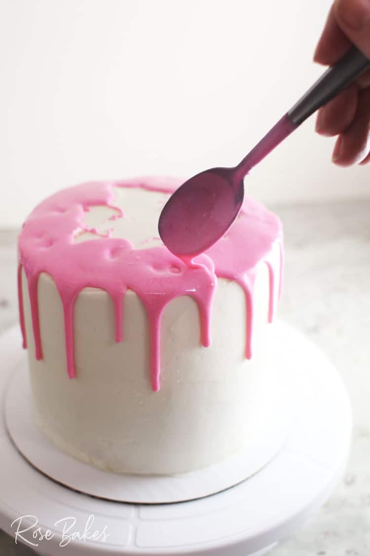 Pink canned frosting being spooned onto the edge of a chilled white cake to create a drip cake
