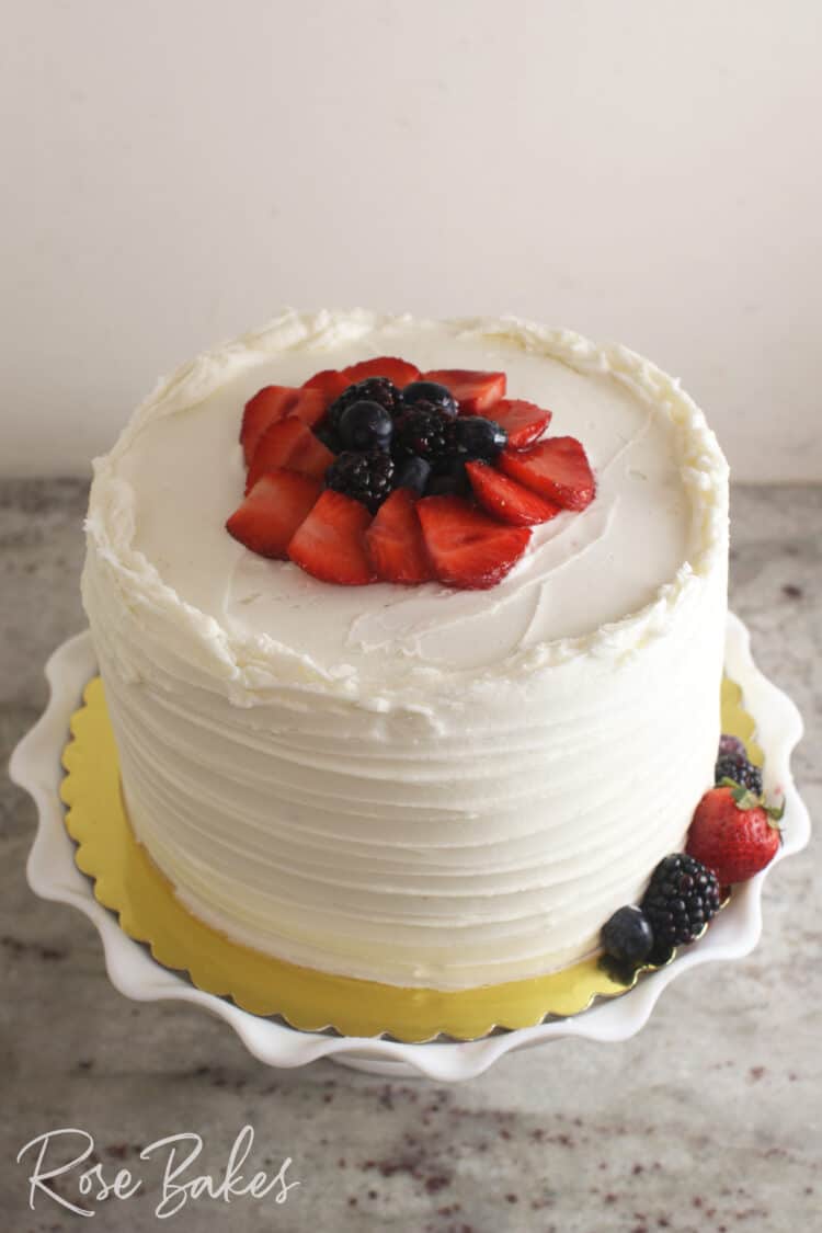 Chantilly Cake with sliced strawberries, blueberries, and blackberries on top. Displayed on a white ruffled cake stand.