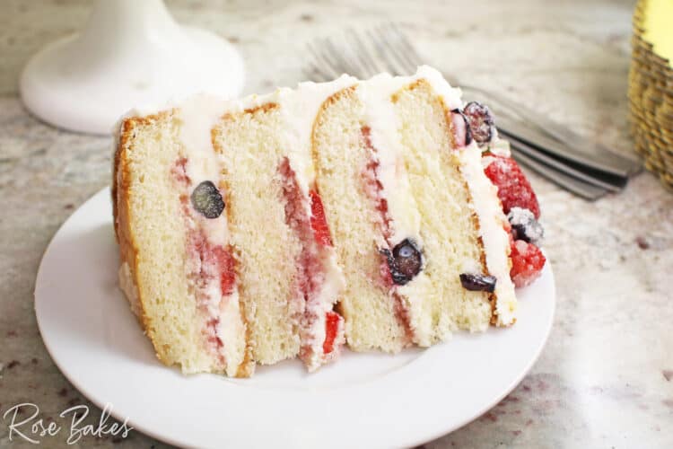 Slice of Chantilly Cake with berries in the filling and on top of the cake.  The cake is on a round white plate.