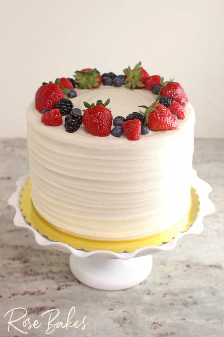 Chantilly Cake with a fresh berry wreath on top. Displayed on a white ruffled cake stand.