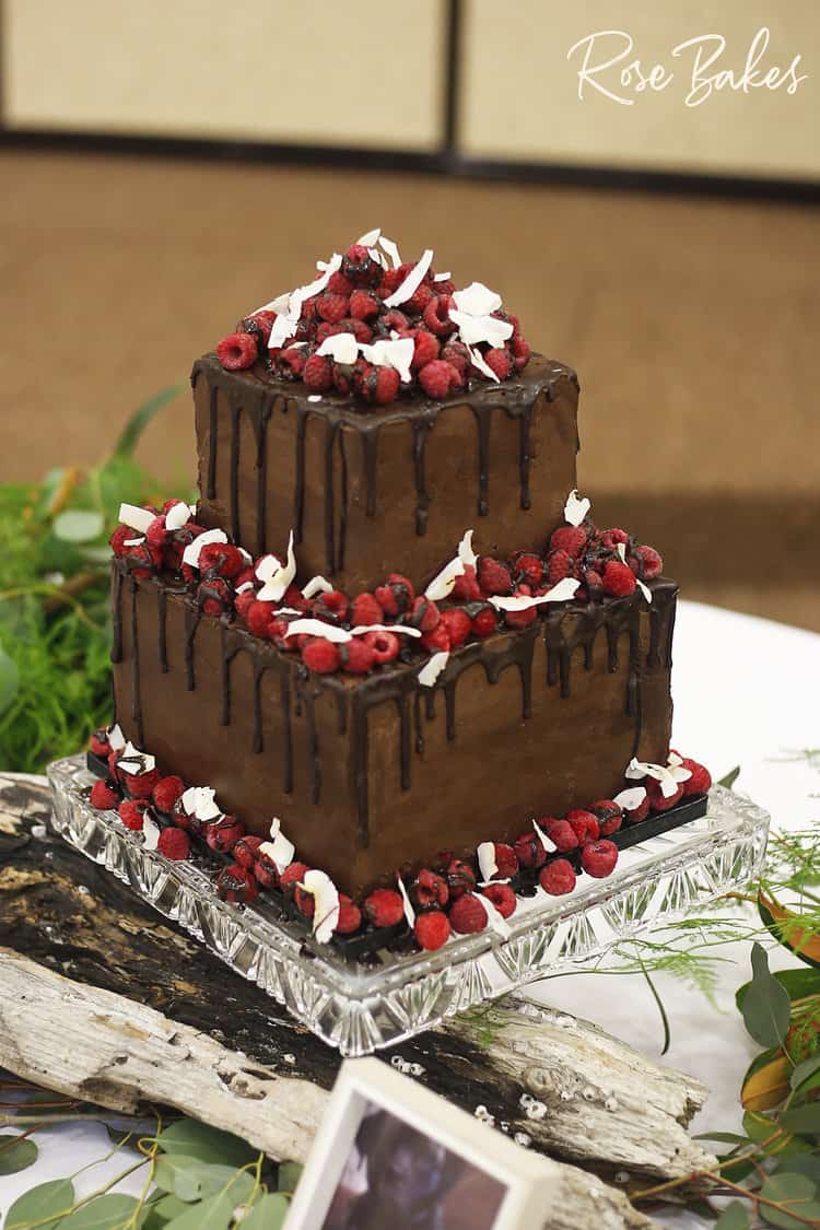 two tiered square chocolate cake with chocolate drip, fresh raspberries, and toasted coconut shavings
