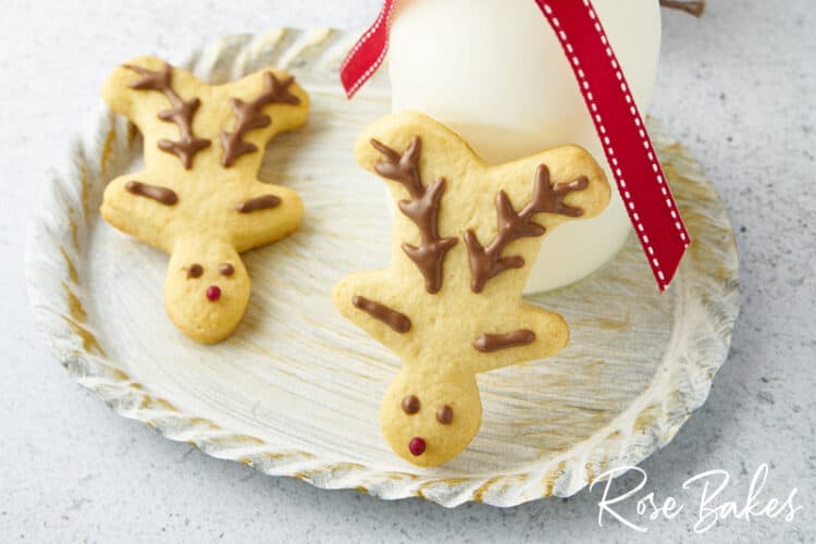 Two simply decorated Christmas reindeer cookies on a white platter with one propped up against a glass of milk.