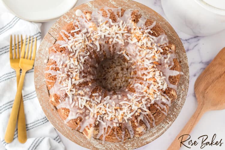 easy one bowl coconut bundt cake on a wooden cake stand. Two gold forks are to the left and a wooden spatula to the right of the cake.