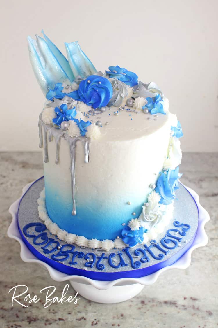 graduation cake with blue ombre spray on white frosting plus blue and white piped decorations and silver drip