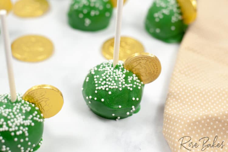 Green cake pops with white nonpareil sprinkles and a gold coins on top. Gold coins are sprinkled on the table among the cake pops.
