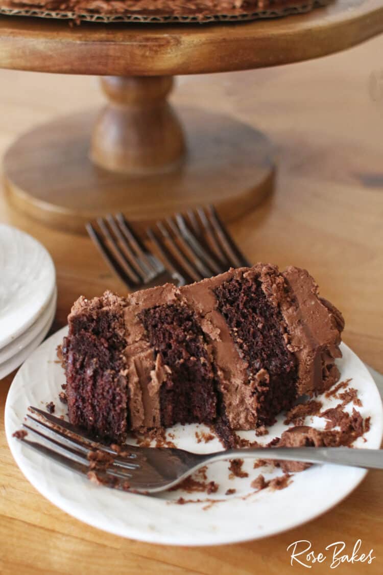 A slice of chocolate cake filled with chocolate frosting on a white plate.  A fork is resting on the plate and a couple of bites have been eaten from the slice of cake.  The wooden cake stand is in the background.