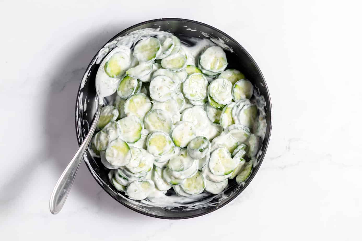sliced cucumbers tossed in german salad dressing in a black bowl with a spoon