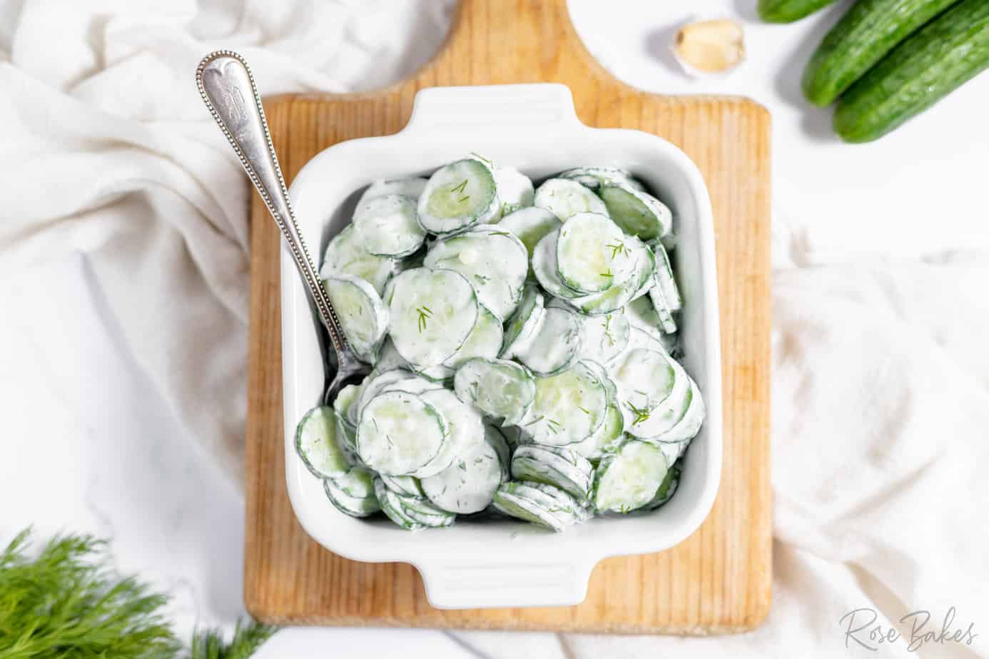 german cucumber salad tossed in a creamy white sour cream dressing in a white bowl with a spoon on a cutting board