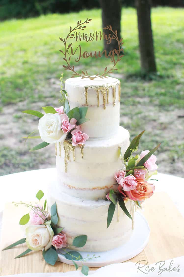 four tiered white wedding cake with gold drip on alternating edges plus fresh pink and white flowers