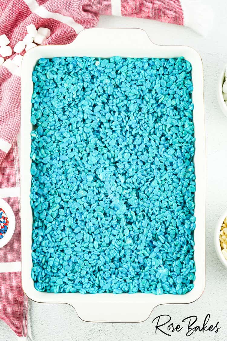 Blue rice krispies pressed flat in a white baking dish on top of white and red rice krispies