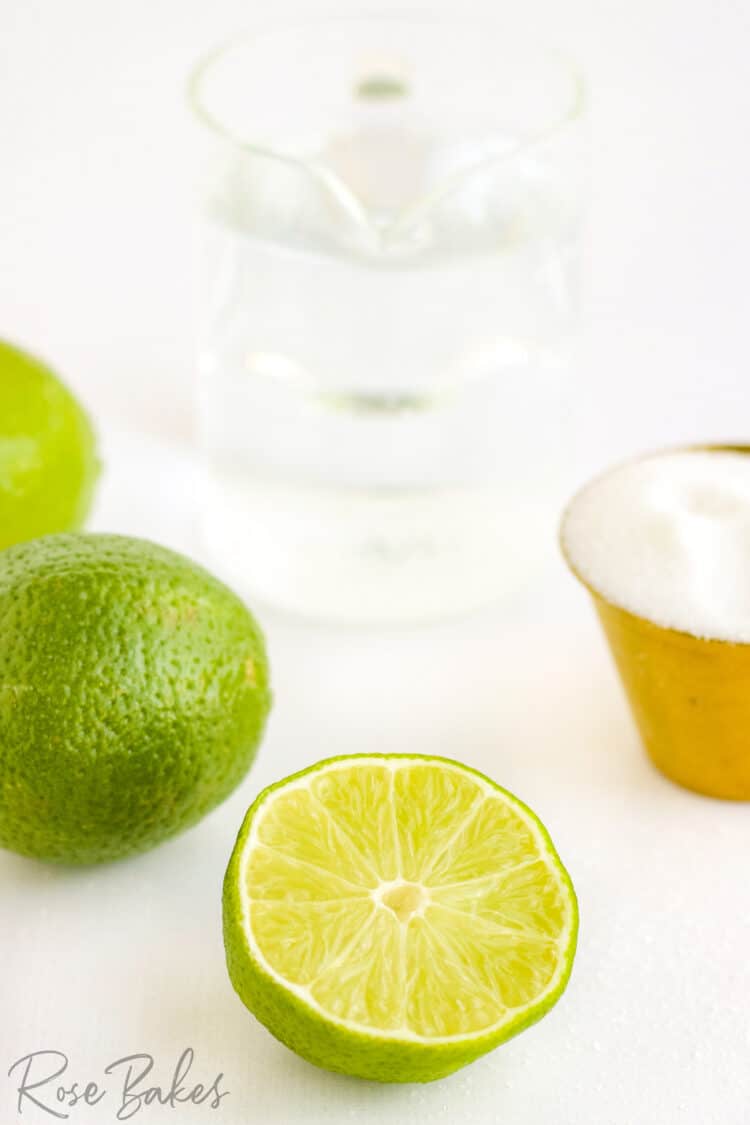 Whole limes, a lime sliced in half, a copper bowl of sugar, and clear pitcher of water.