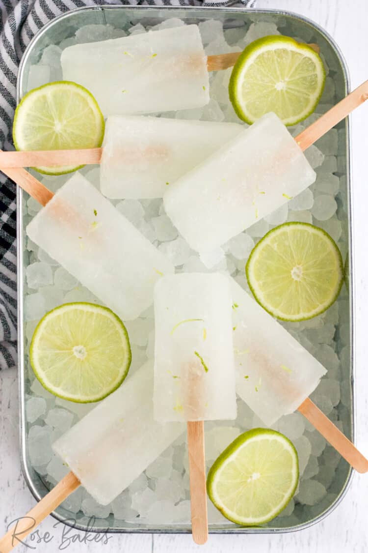 Popsicles with wooden popsicle sticks laying in a dish of crushed ice and slices of lime.