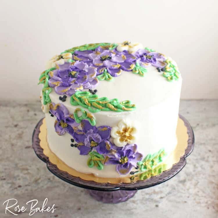 purple and green buttercream painted flowers and leaves with edible gold trim on white buttercream one tier cake on glass cake stand 