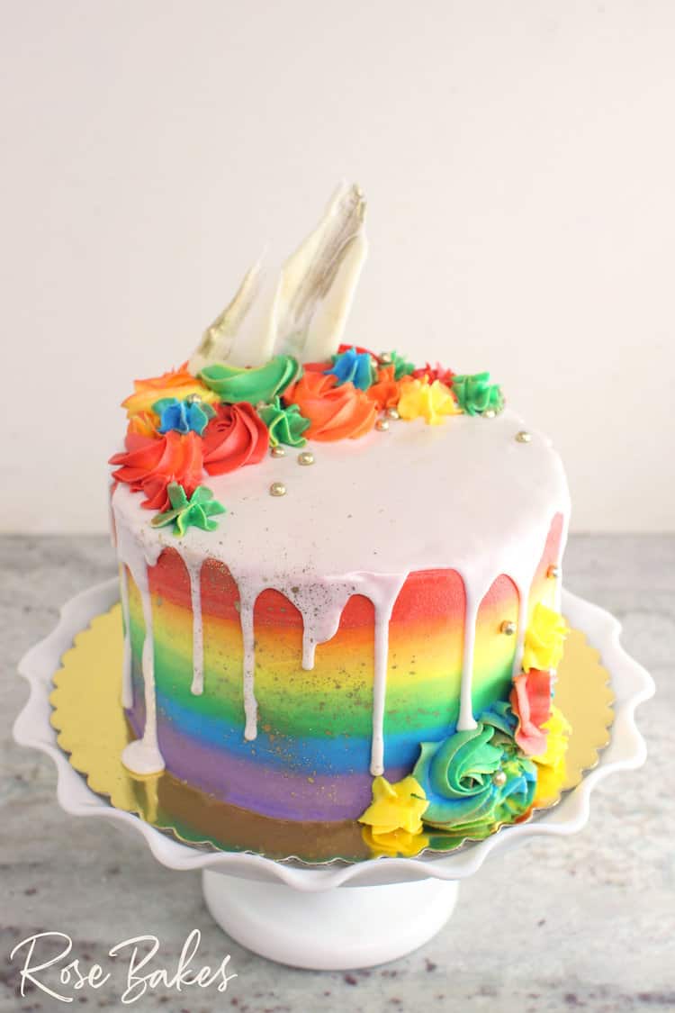 rainbow buttercream cake with white drip and rainbow piping plus white chocolate brushes and gold splatter