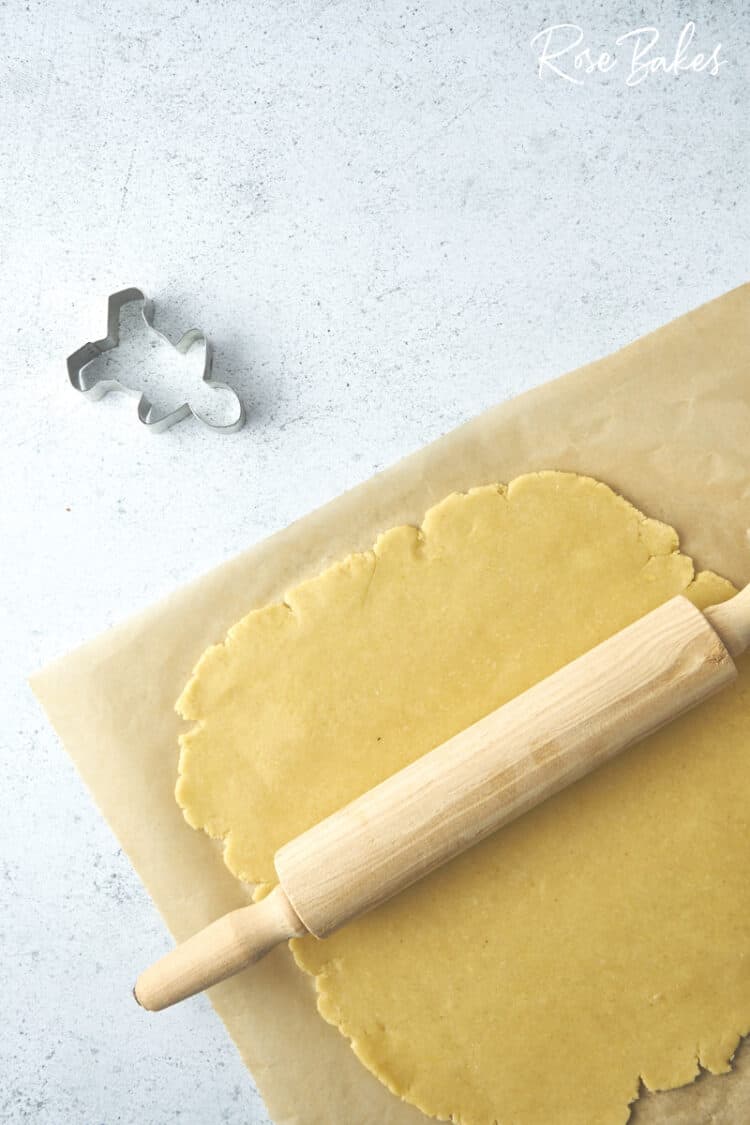 the cookie dough rolled out on a sheet of parchment.  A rolling pin sits on top of the dough and a gingerbread man cookie cutter is off to the side.
