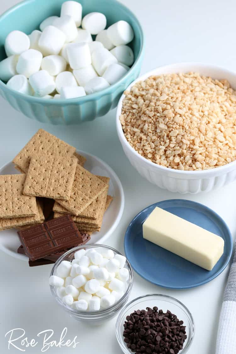 smore rice krispie treats ingredients on a counter including rice krispies cereal in a white bowl, a plate of graham crackers and chocolate bars, a stick of butter on a blue plate, two bowls of marshmallows and a small bowl of mini chocolate chips