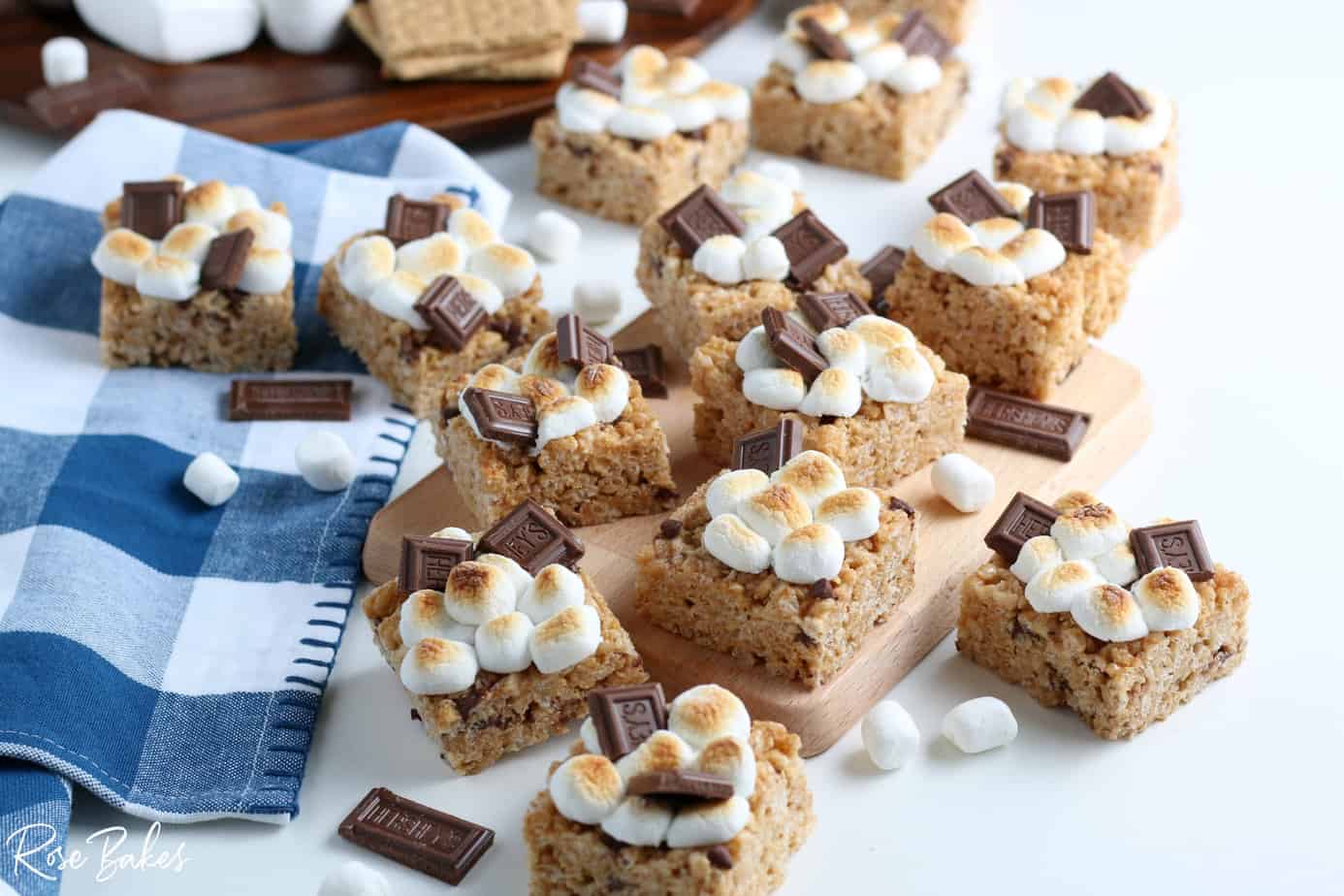 Smores Rice Krispie Treats cut into squares and spread on a cutting board and checkered kitchen towel. They are topped with toasted mini marshmallows and a piece of chocolate.