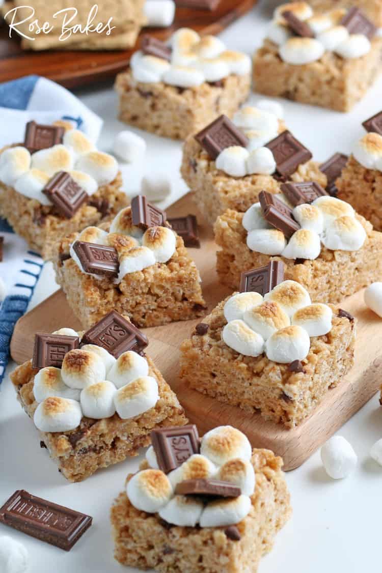 S'mores Rice Krispie Treats cut into squares and spread on a cutting board and checkered kitchen towel. They are topped with toasted mini marshmallows and a piece of chocolate.
