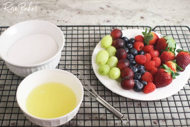 Ingredients for making sugared berries. Top left: bowl of sugar, bottom left: pasteurized egg whites and whisk, right: plate of green grapes, purple grapes, blueberries, raspberries, and strawberries