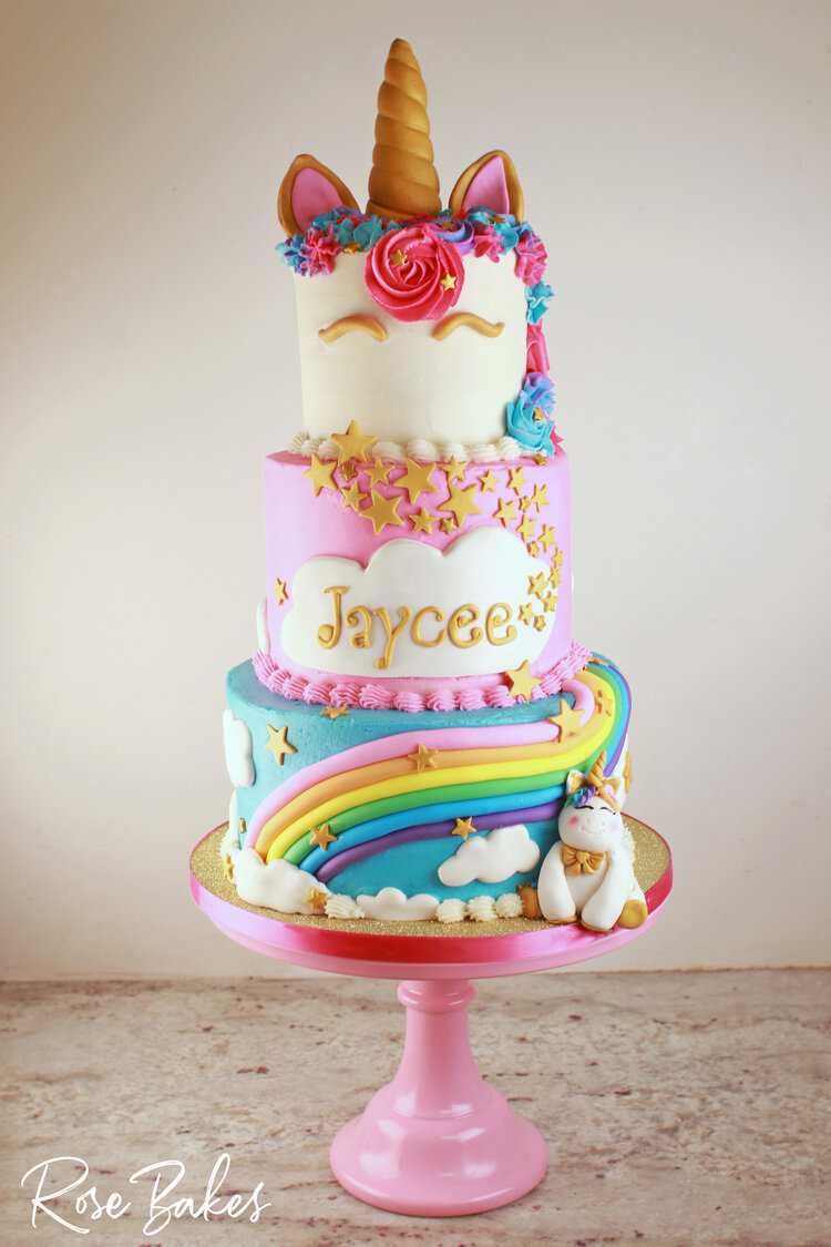 Three tiered cake with the top tier topped with a unicorn horn and ears, buttercream mane, and fondant eyes. Pink middle tier with a large cloud with the name Jaycee and a cascade of stars. The bottom tier is blue with a fondant rainbow with a fondant unicorn figure sitting next to it on a gold glittery cake board. 