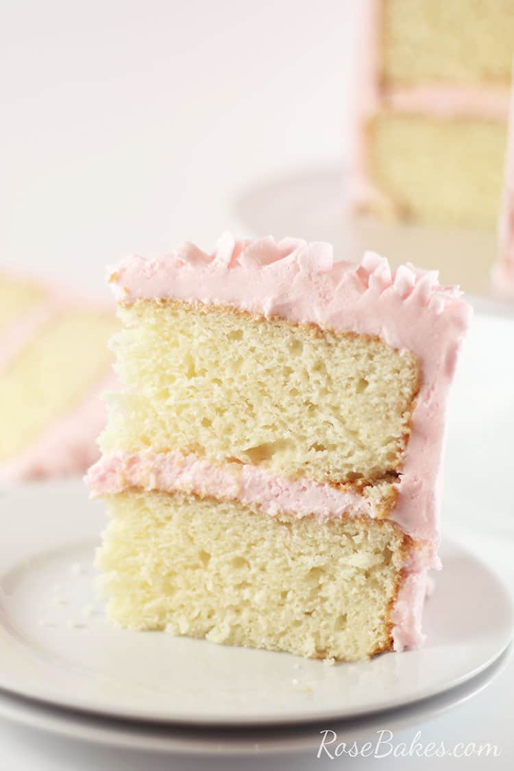 A slice of White Almond Sour Cream Cake with light pink frosting standing up on a white plate.