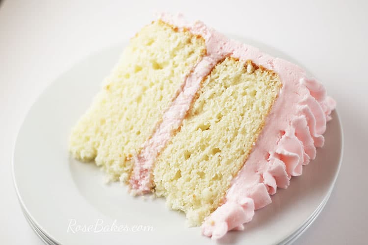 A slice of White Almond Sour Cream Cake with light pink frosting laying on a white plate.